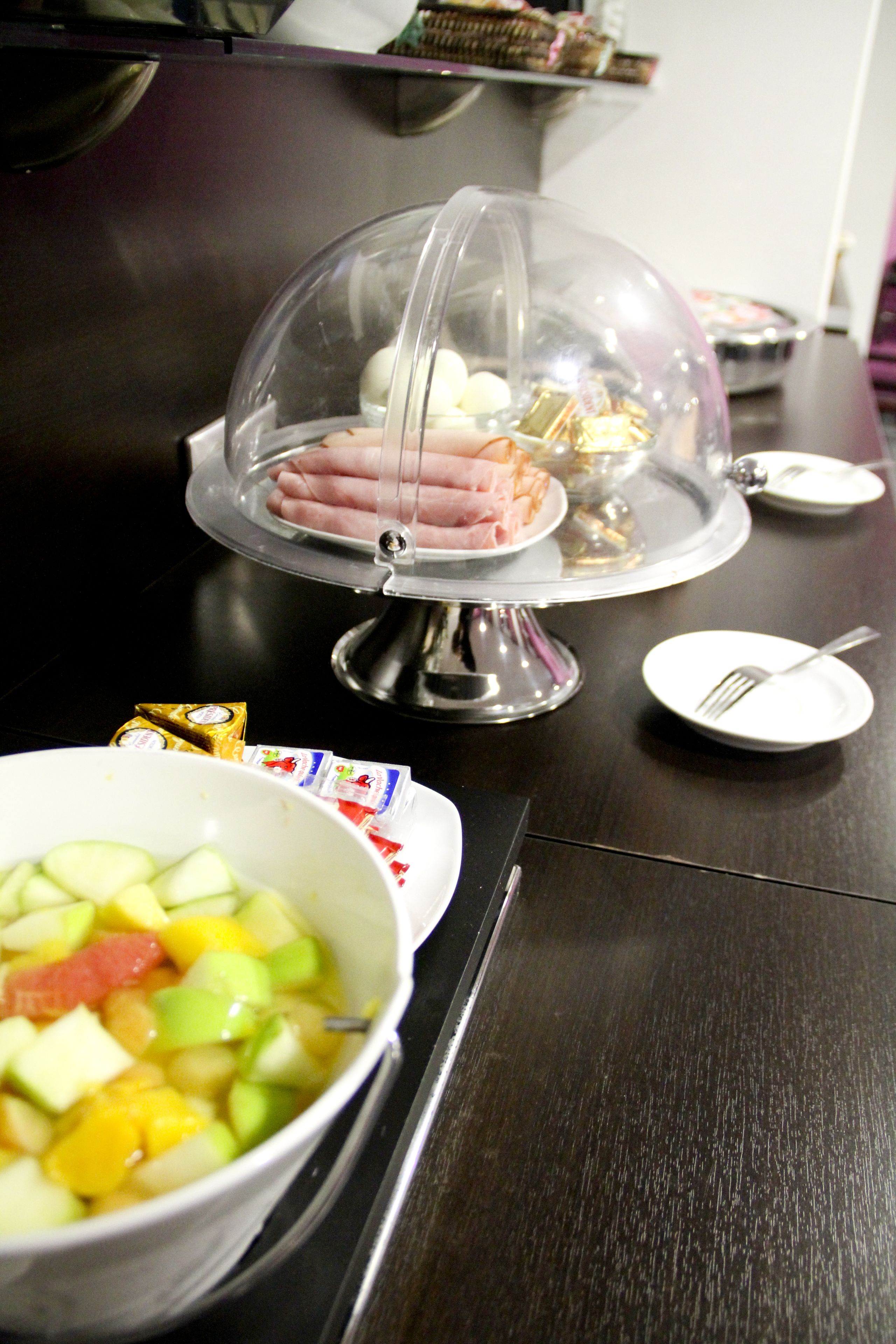 A buffet setup with a covered ham dish, a bowl of fruit salad, and a small plate with a fork.