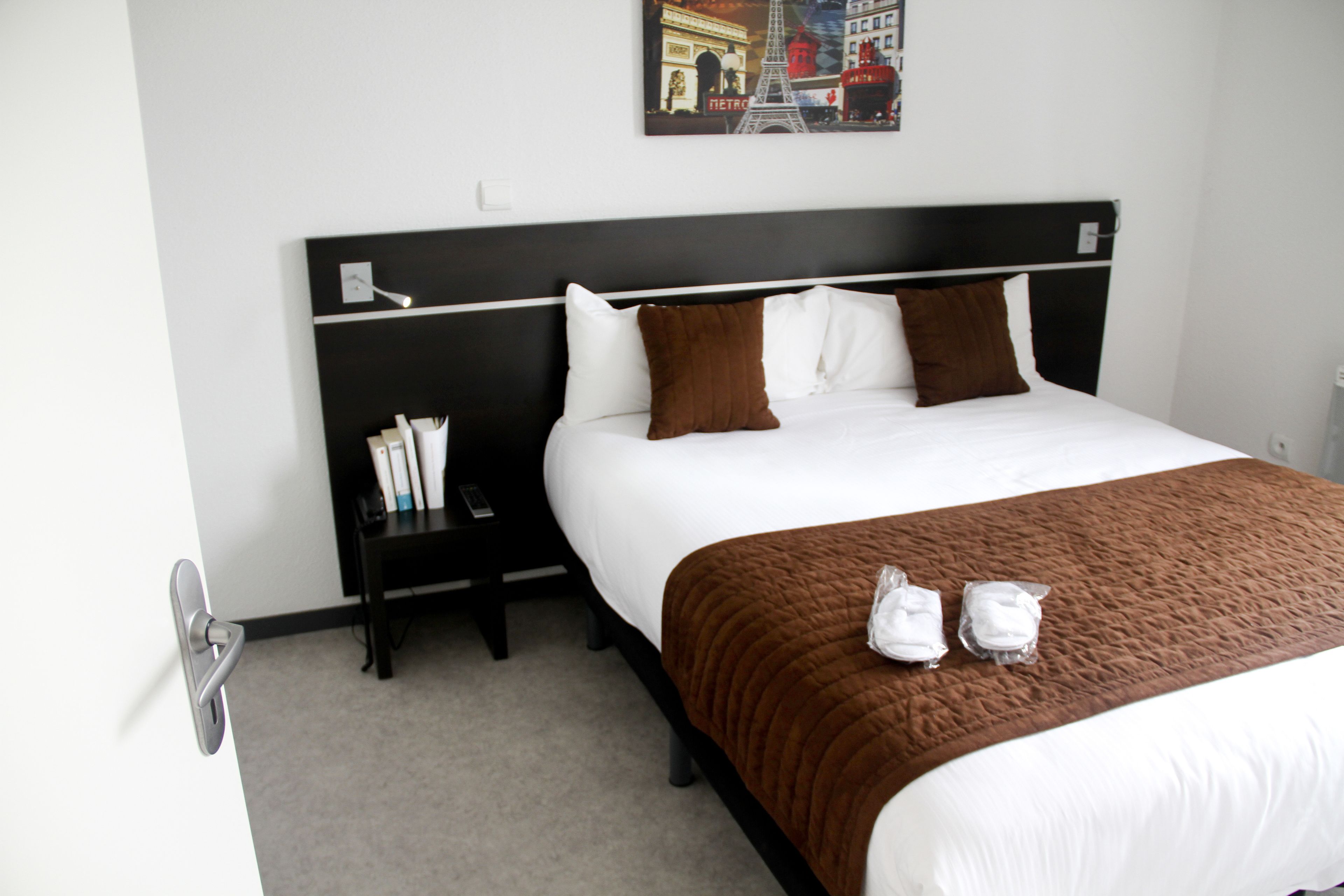 A hotel room with a large bed, a nightstand with a phone and a lamp, and a painting of the Eiffel Tower on the wall.