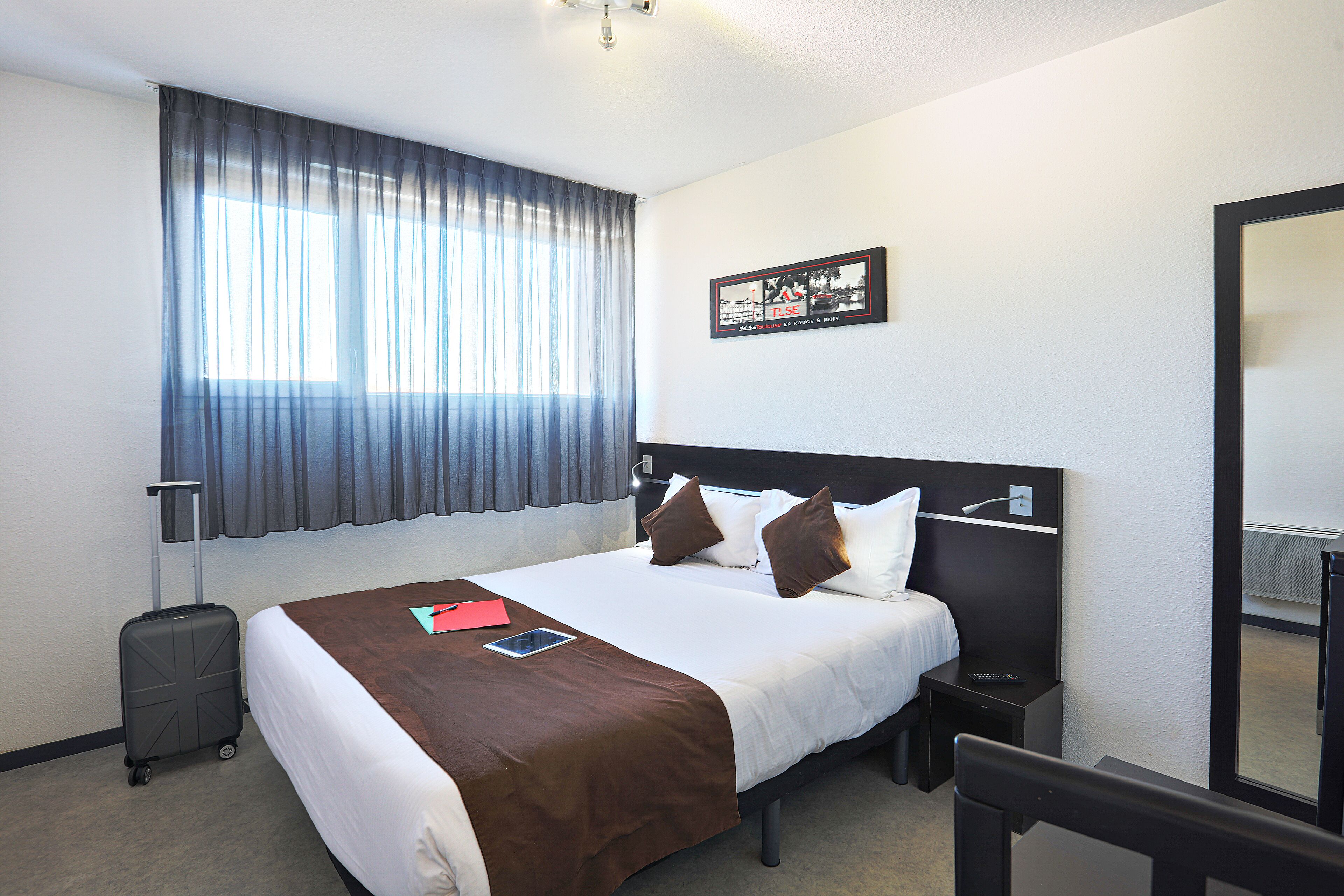 Une chambre d’hôtel avec un grand lit, une table de chevet, une valise, une fenêtre avec des rideaux et un tableau accroché au mur.
