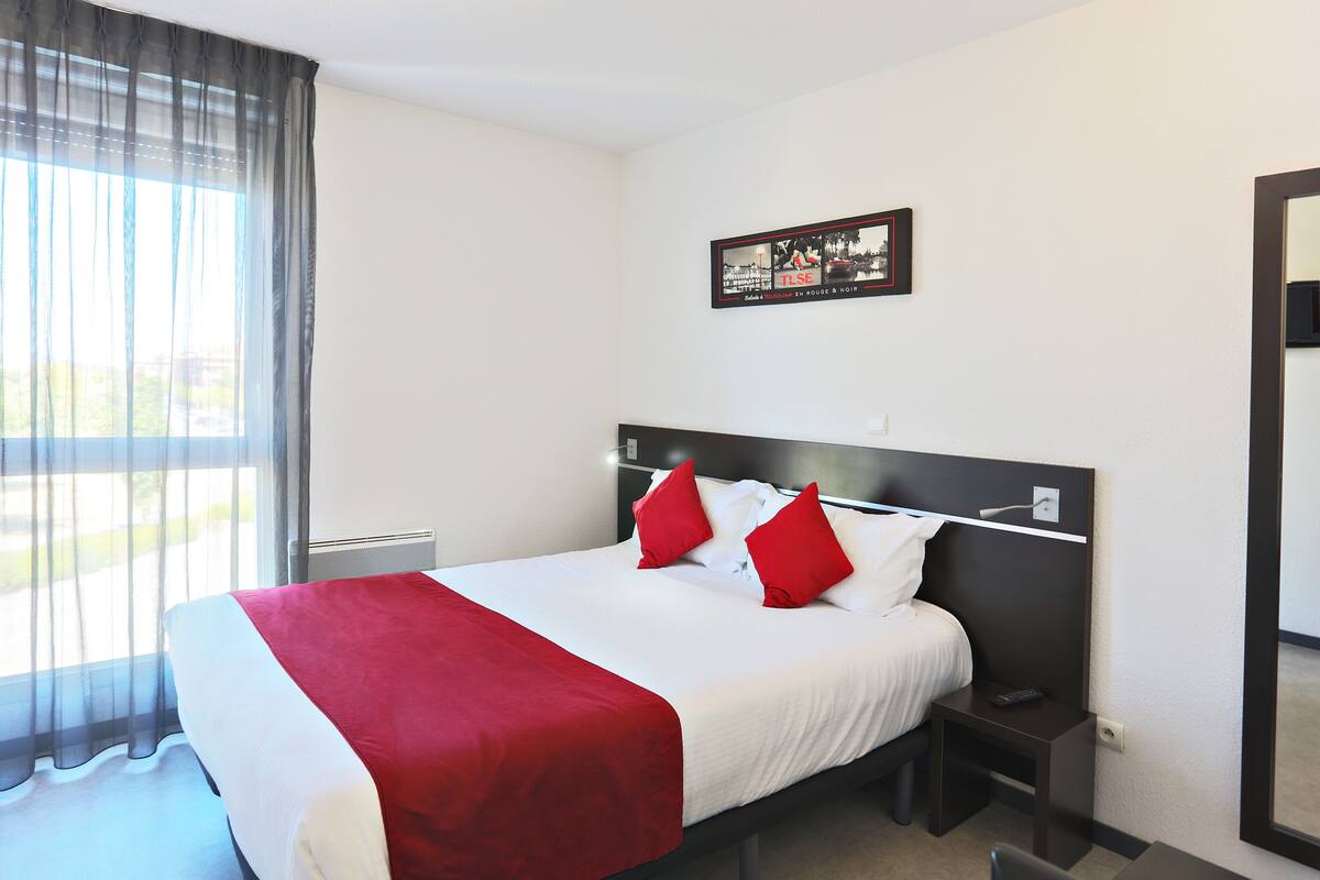 Une chambre d’hôtel avec un lit, une table de chevet, un miroir et une fenêtre avec des rideaux.