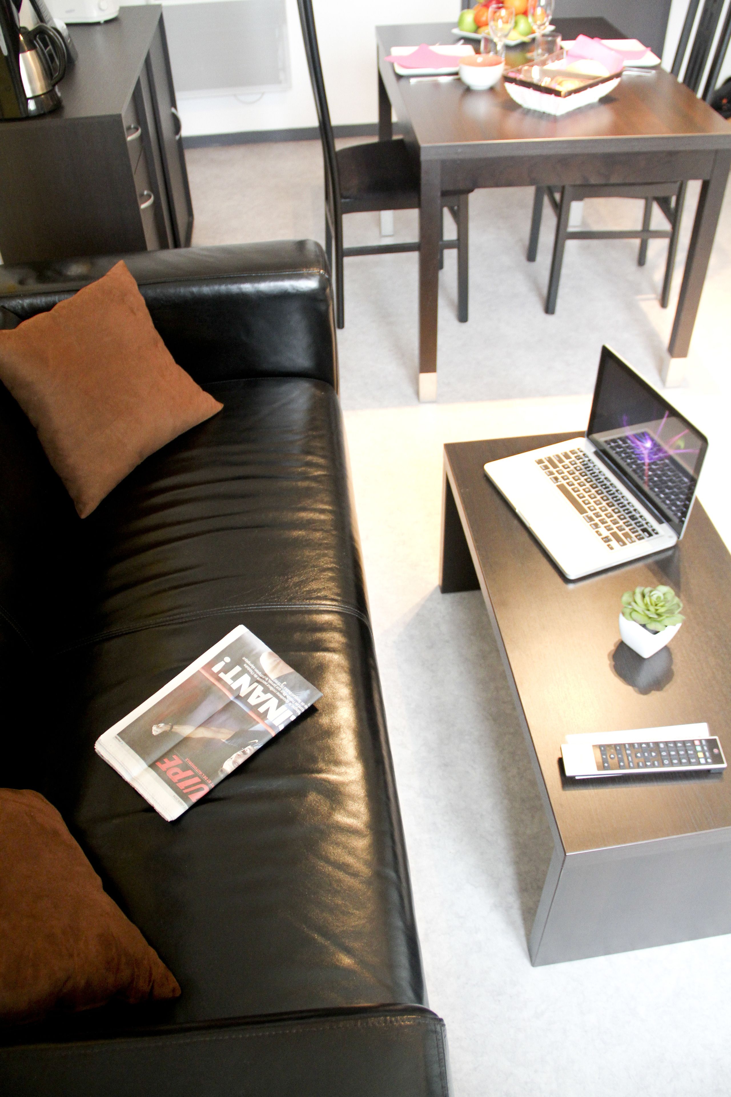 A modern living room with a black leather sofa, a wooden coffee table with a laptop and a small plant, and a dining area in the background.