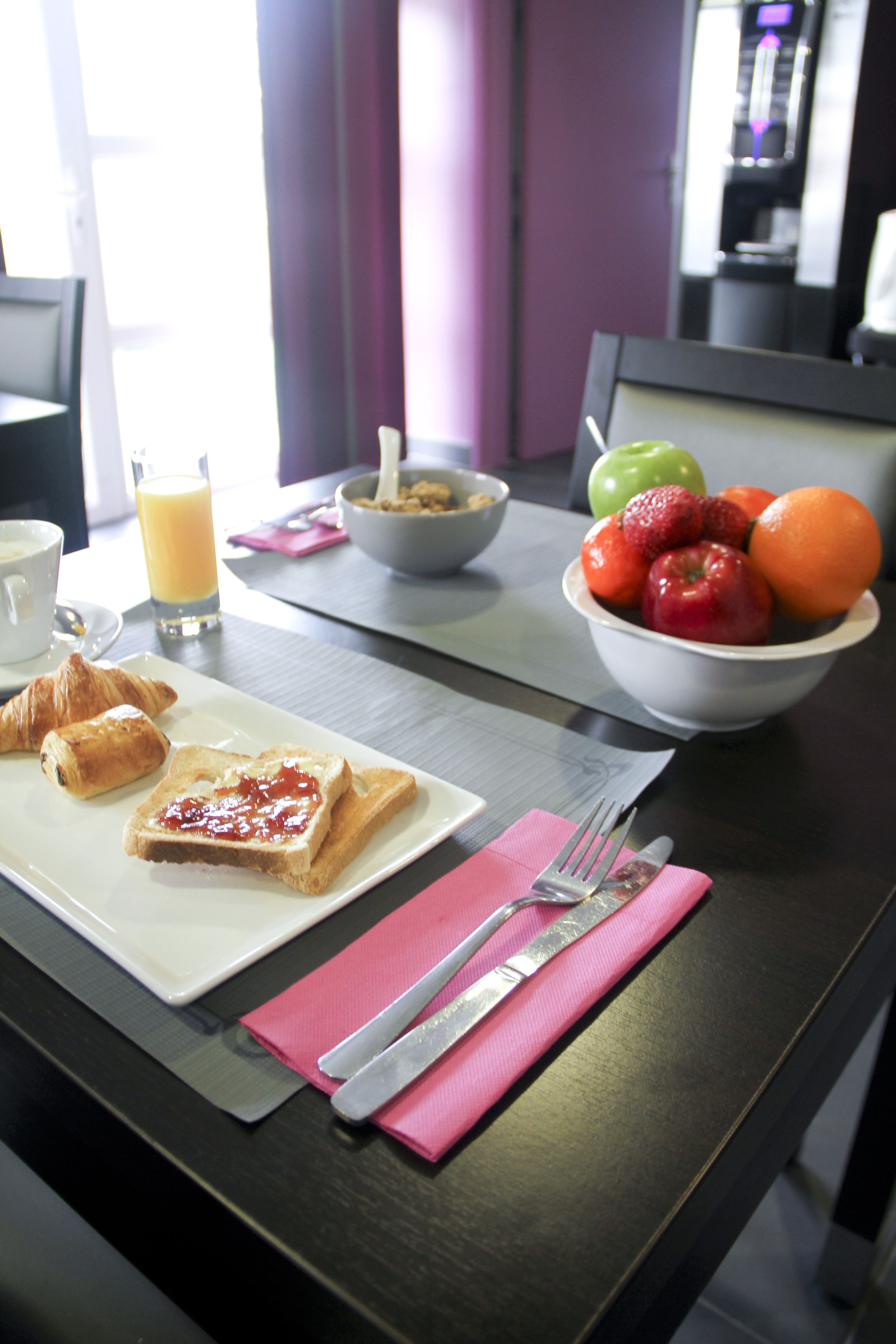 Une table de petit-déjeuner avec des croissants, des tartines de confiture, un bol de fruits, un verre de jus d’orange et une tasse de café.