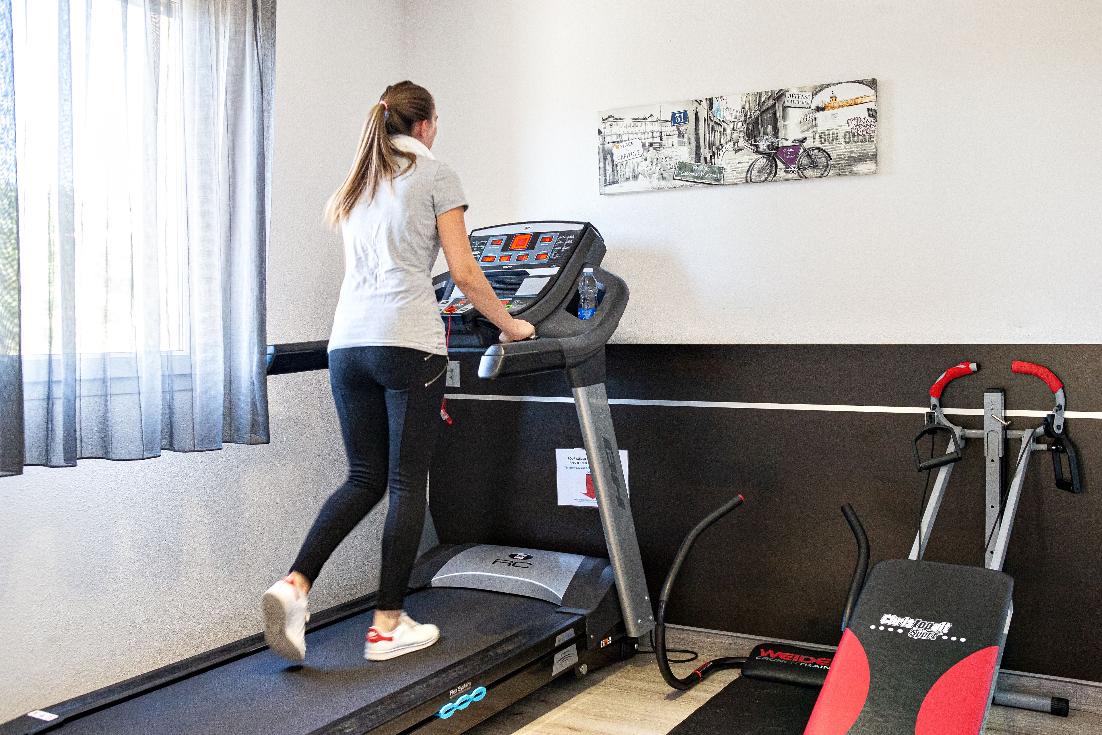 A woman using a treadmill in a room with a window, a wall-mounted picture, and a nearby stationary bike.