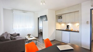 Ein modernes Wohnzimmer mit einer Couch, einem Esstisch und leuchtend orangefarbenen Stühlen. Es gibt eine Küchenzeile mit einer Mikrowelle und einer Kaffeemaschine.