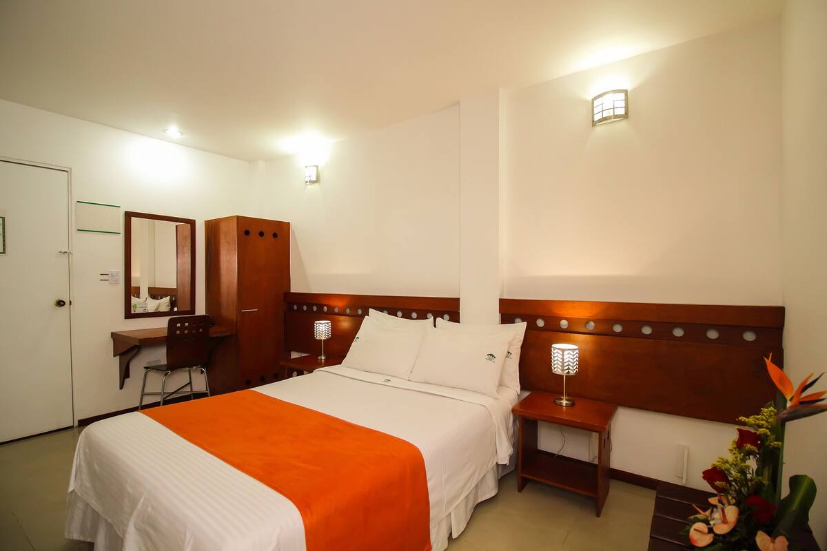 A hotel room with a bed, a desk, a chair, a mirror, a nightstand with a lamp, and a wall-mounted light fixture.