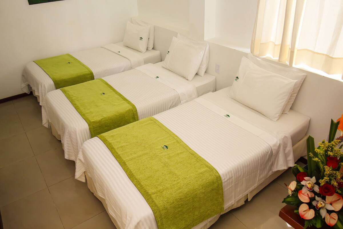 A hotel room with four single beds, each with a white mattress and green bedspread. There is a floral arrangement on the right side of the room.