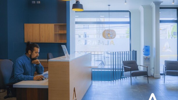 Un área de recepción moderna con un escritorio de madera, una computadora portátil y una persona trabajando. Hay dos sillas, un dispensador de agua azul y un gran ventanal con vistas a edificios.