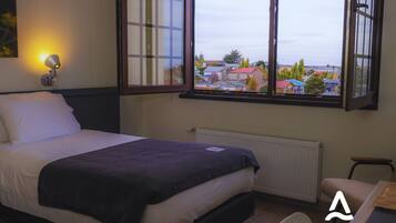 A bedroom with a bed, a desk, and a window with a view of buildings.