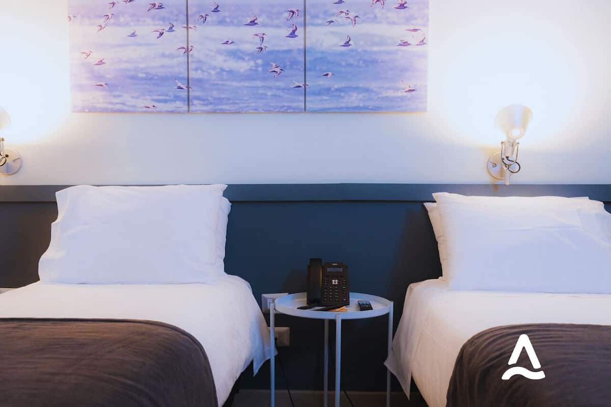 A hotel room with two beds, a small table with a phone, and a wall with a bird painting.