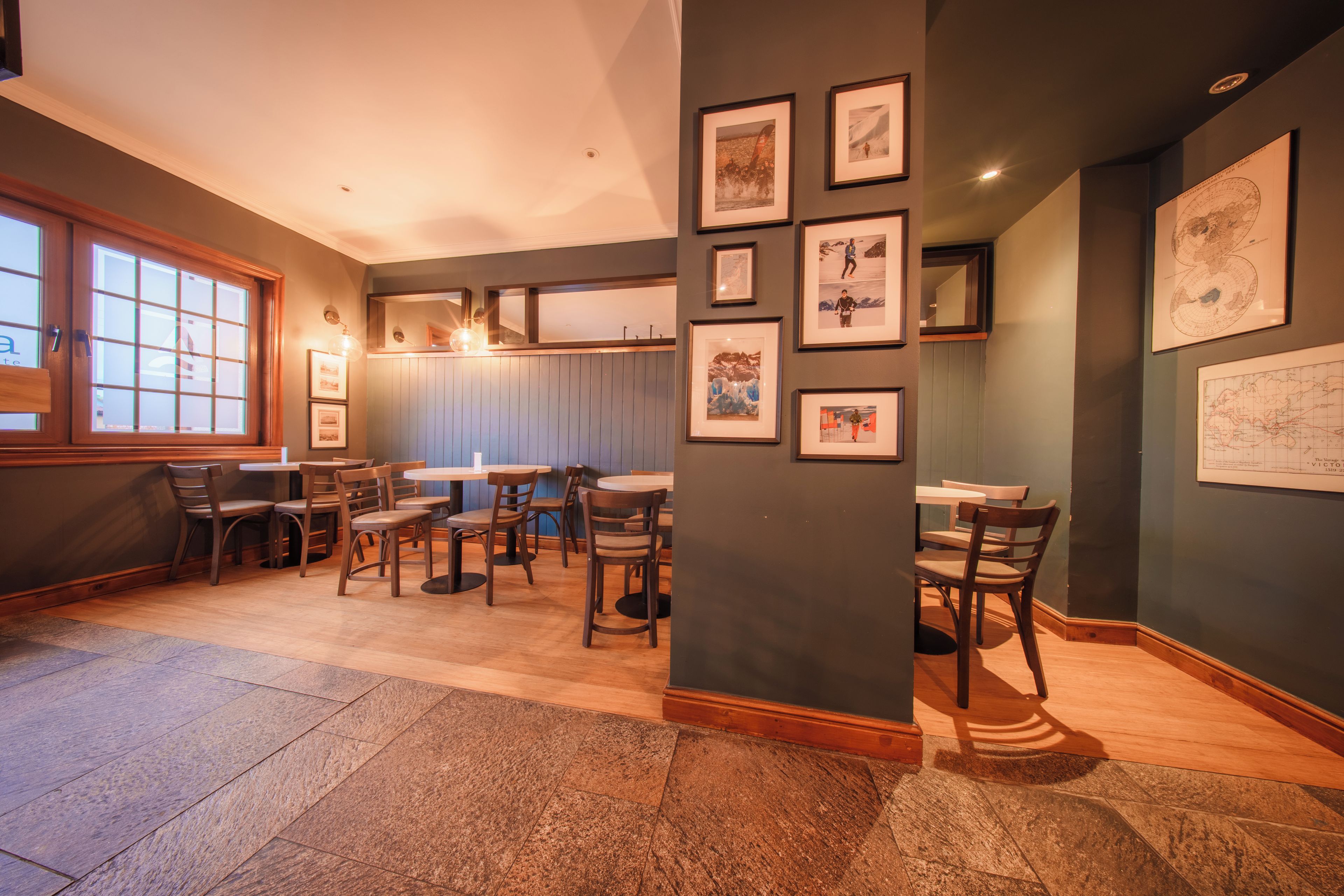 Interior de restaurante con mesas y sillas de madera, cuadros en las paredes y un ventanal grande a la izquierda.