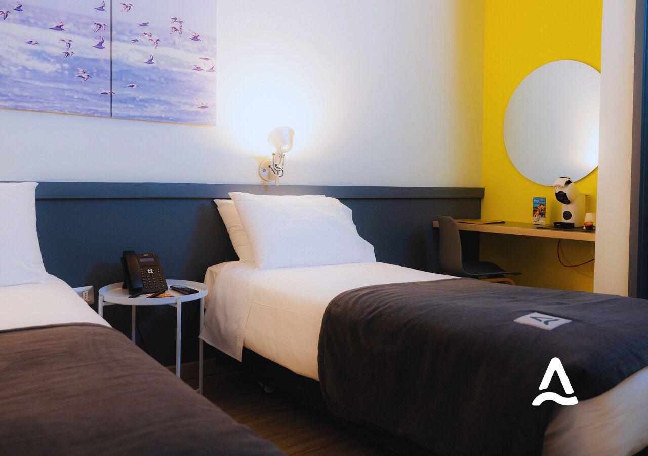 Une chambre d’hôtel avec deux lits, une table de chevet avec un téléphone, une lampe fixée au mur et un bureau avec un ordinateur et un miroir.