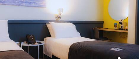 Habitación de hotel con dos camas, una mesita de noche con teléfono, una lámpara de pared y un escritorio con computadora y espejo.