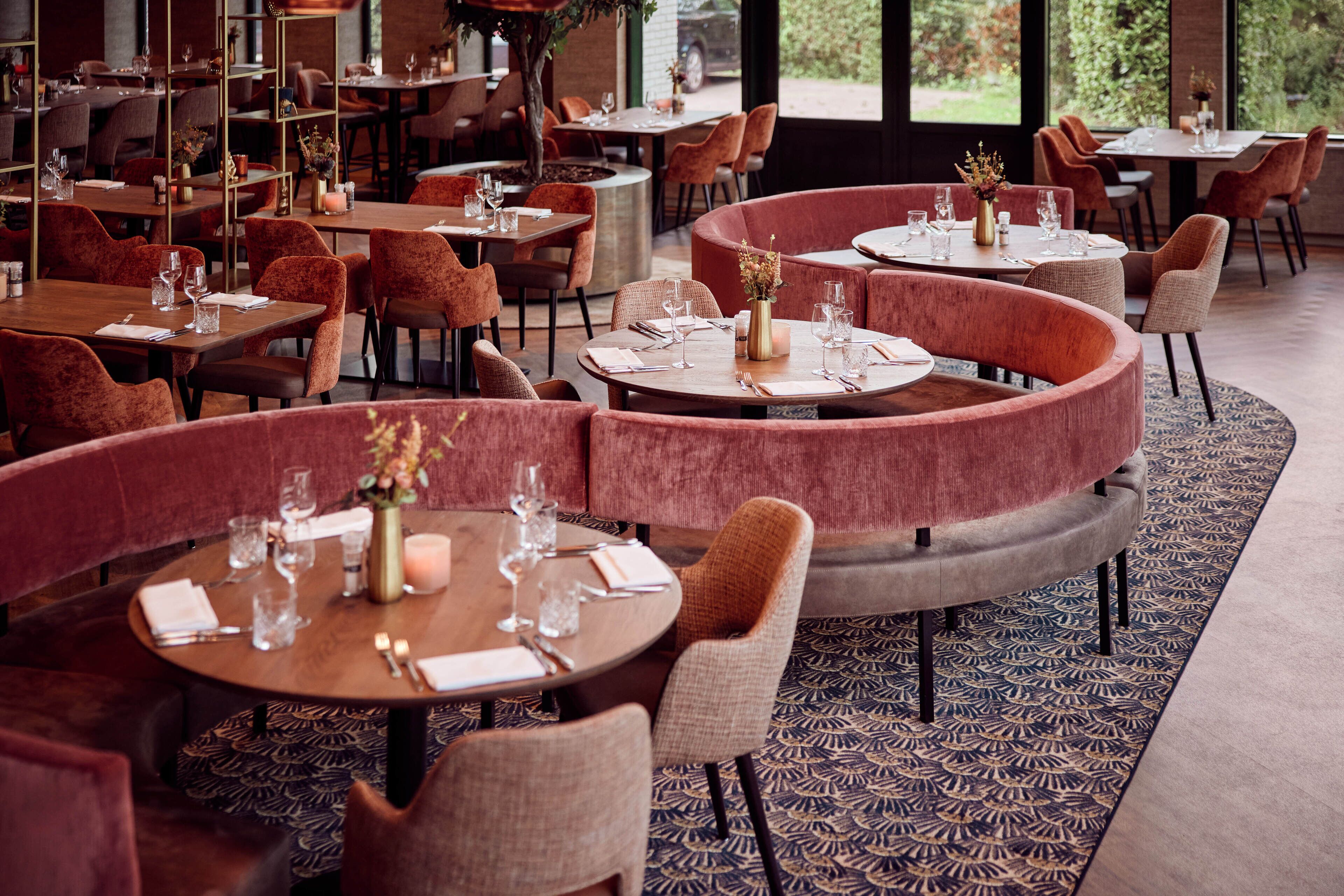Un restaurante con mesas redondas y asientos acolchados, que presenta un suelo estampado y grandes ventanales.