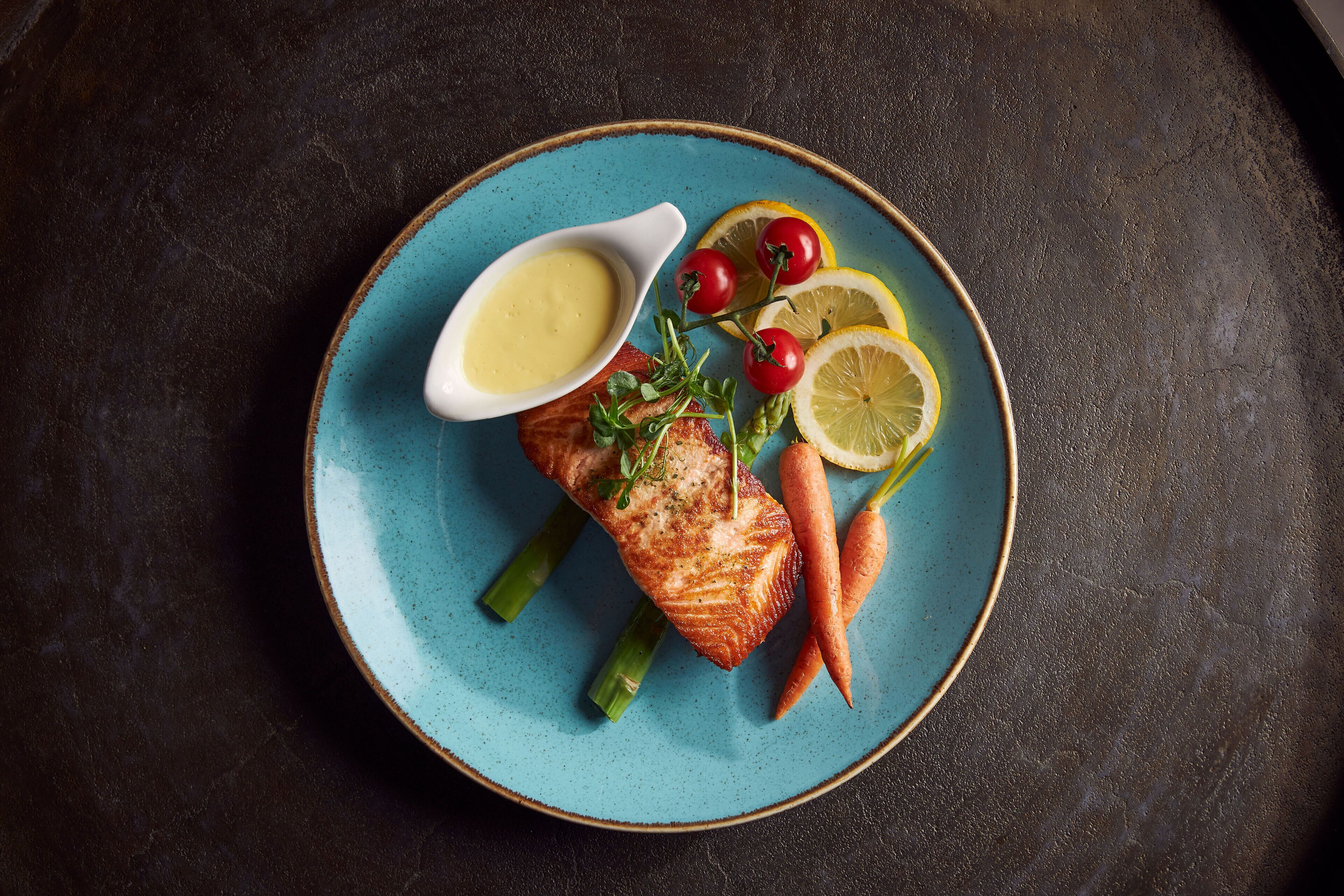 Een bord met een stuk gegrilde zalm, partjes citroen, kerstomaatjes en asperges op een blauw bord.