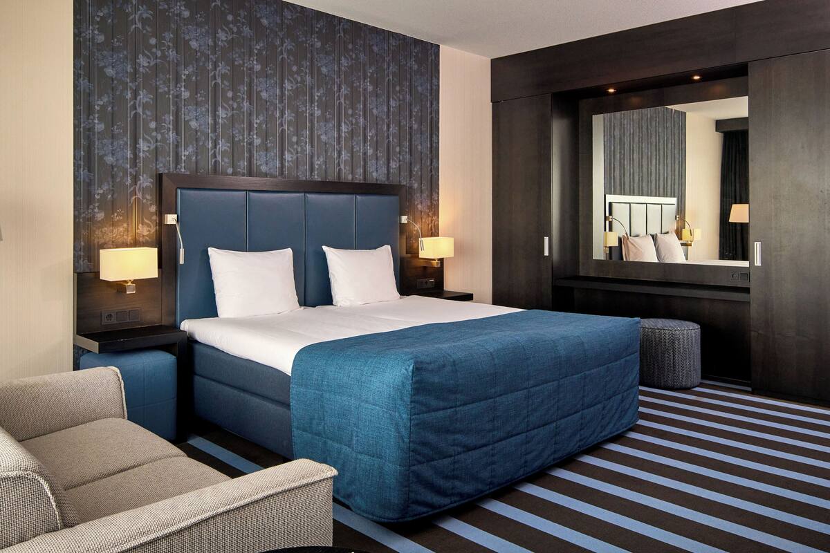 A hotel room with a large bed, two bedside tables with lamps, a chair, a small ottoman, and a large mirror.