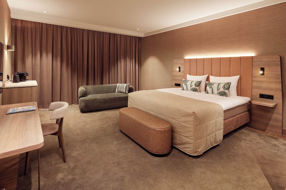 A modern hotel room with a large bed, a sofa, a desk with a chair, and a small table.