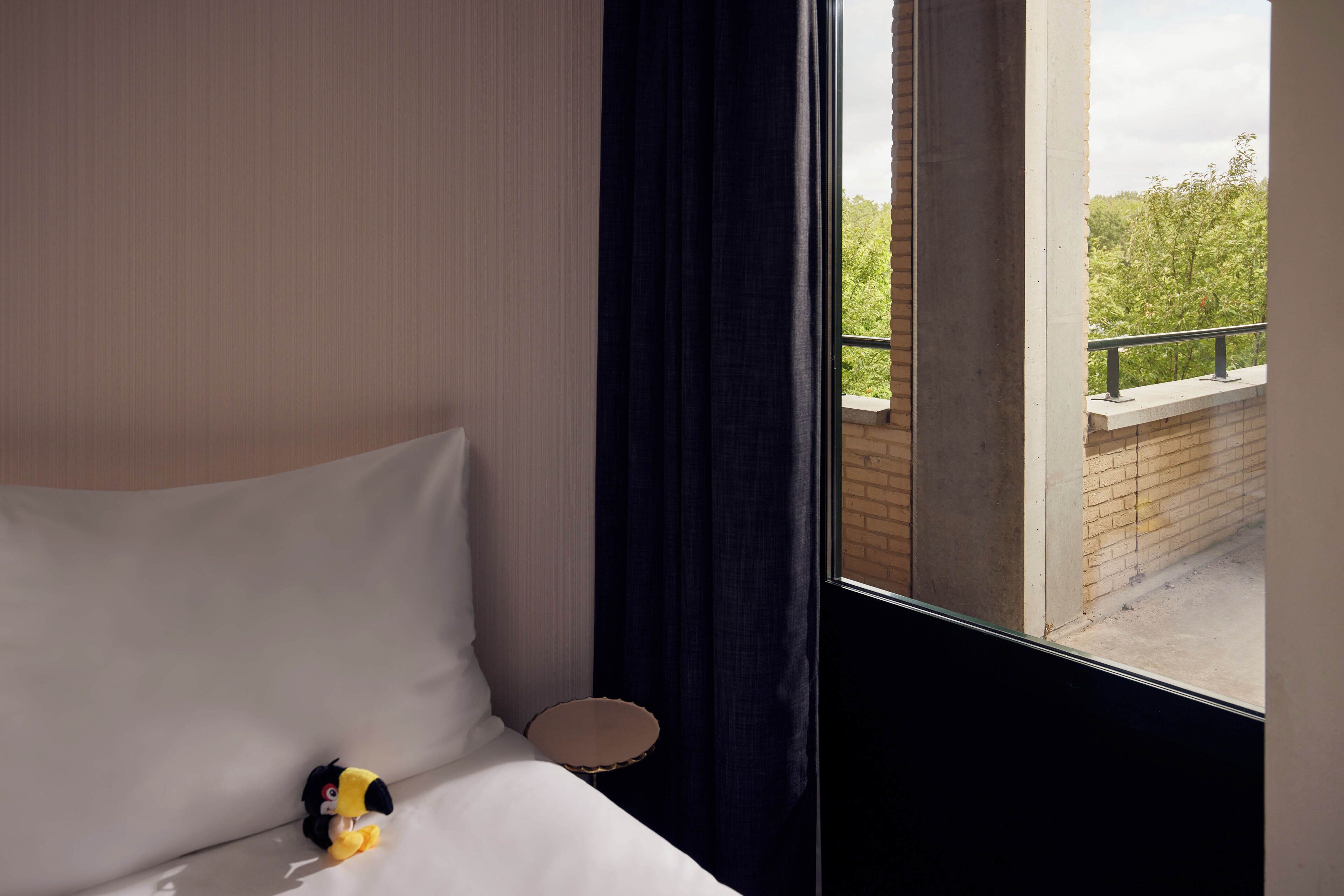 Quarto de hotel com cama, uma mesinha redonda e vista para um muro de tijolos e vegetação lá fora.