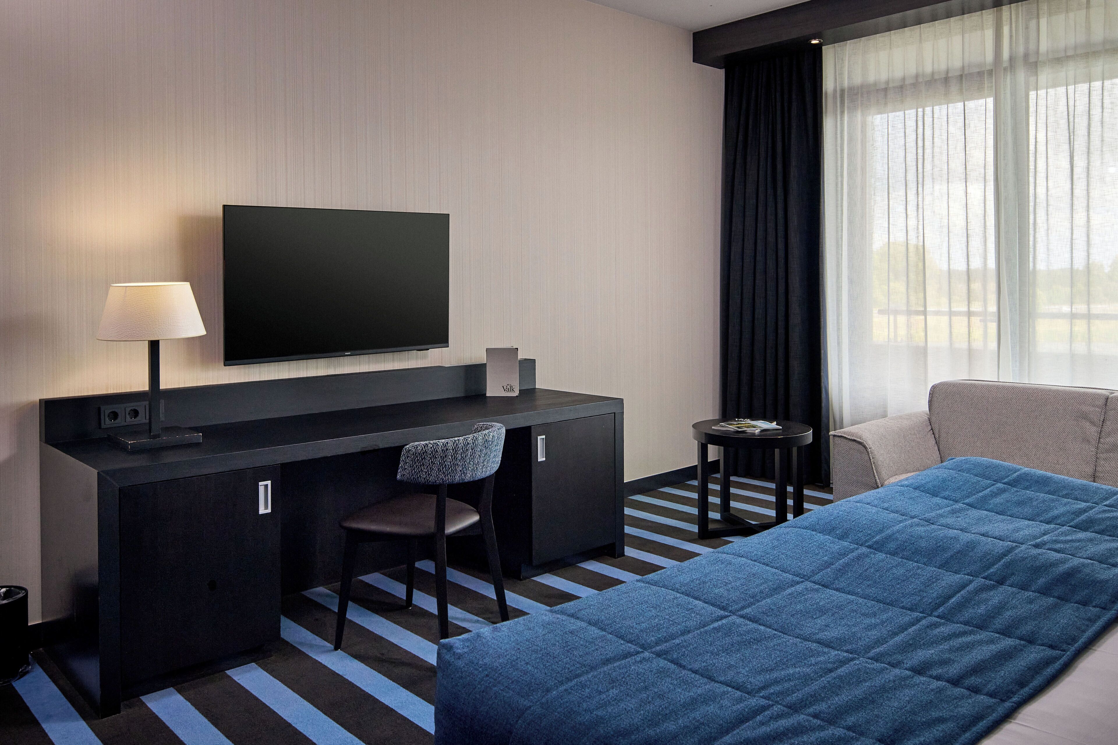 Camera d'albergo con un letto, una scrivania con una sedia, una televisione e una finestra con tende.