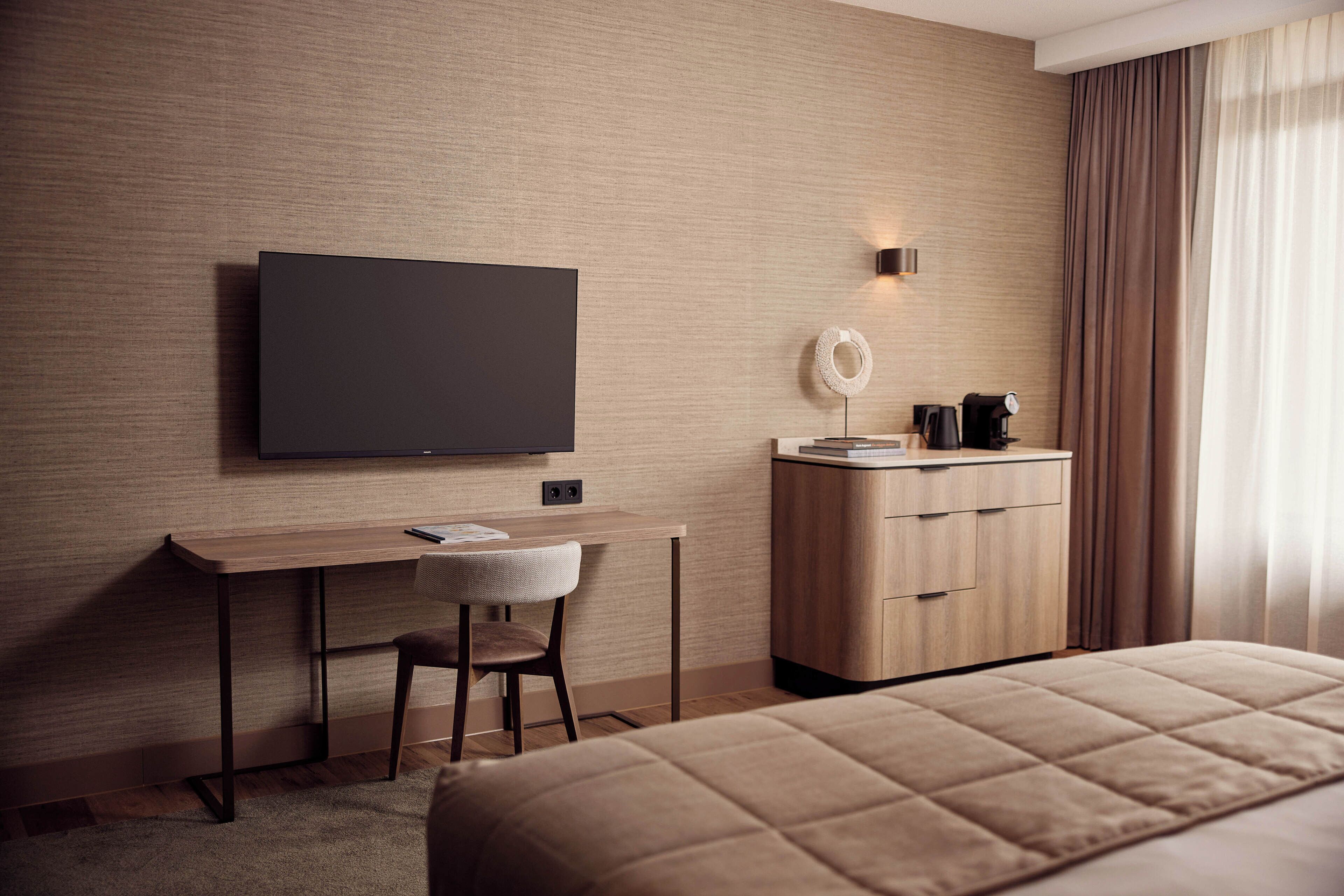 A modern hotel room with a flat-screen TV mounted on the wall, a wooden desk with a chair, a bedside table with a lamp, and a large bed.