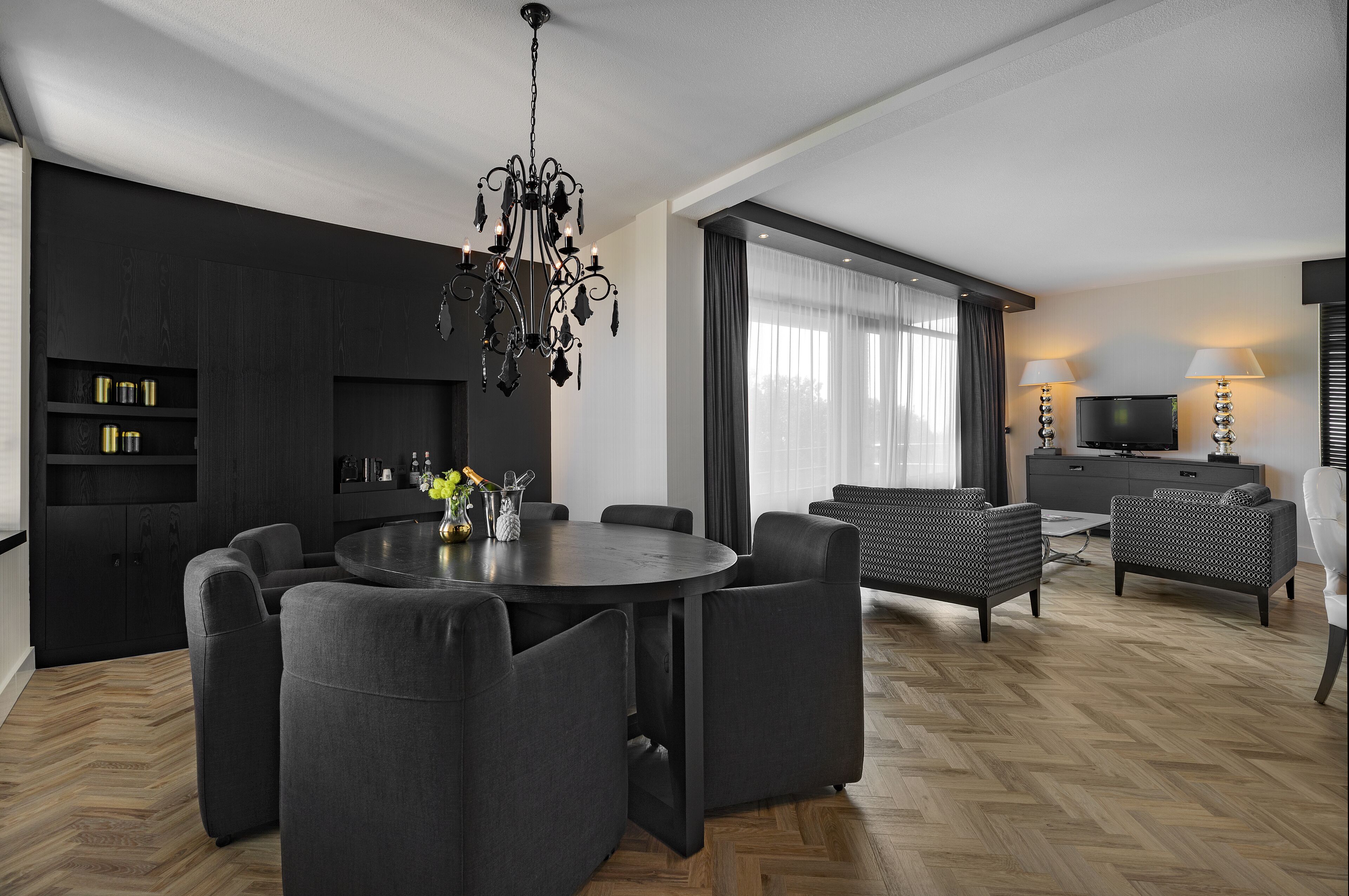 A modern living room with a round dining table, dark sofas, a chandelier, and a TV.