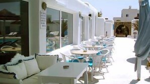 A sunlit outdoor seating area with white tables and chairs, a white sofa, and a view of the sea.