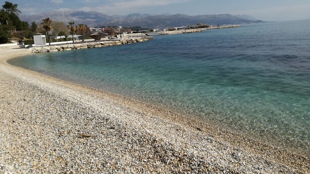 A pebble beach with clear water, a rocky shoreline, and a distant mountain range. - Hotel More (Split)