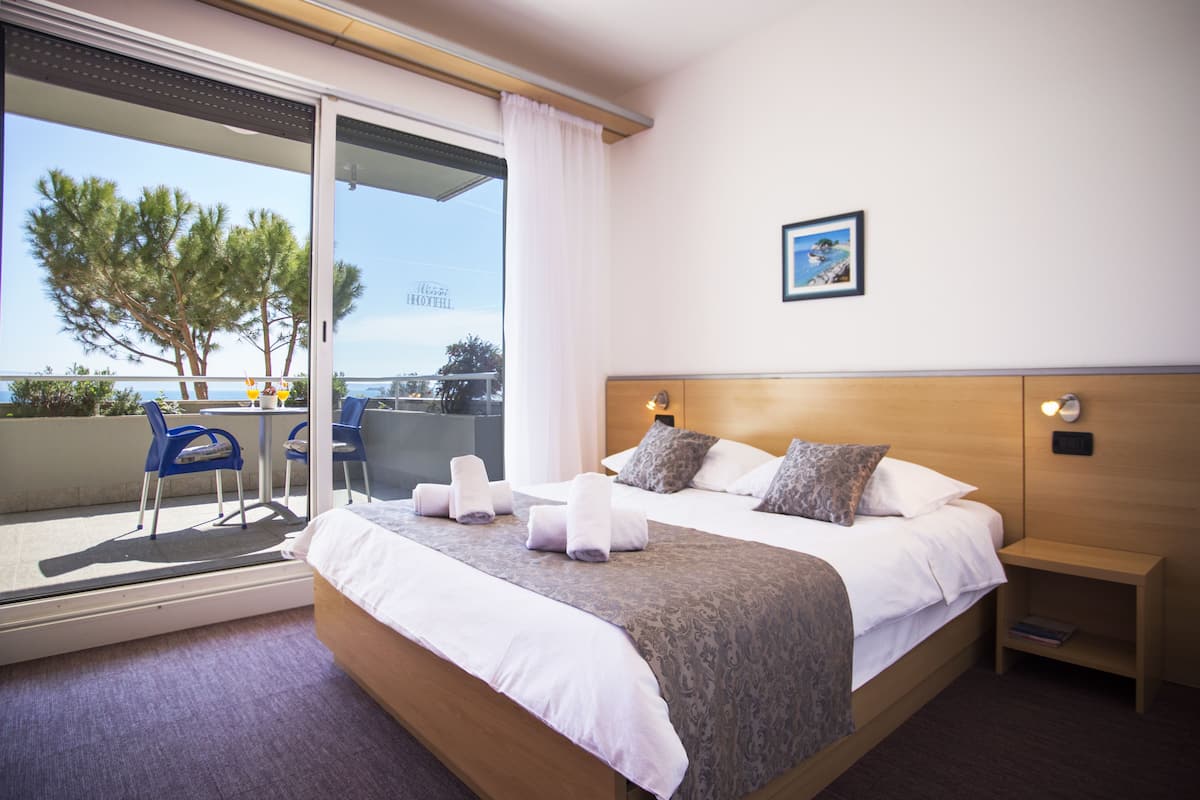 A hotel room with a bed, a bedside table, a nightstand, a window with a view of trees and a building, and a chair outside the window.