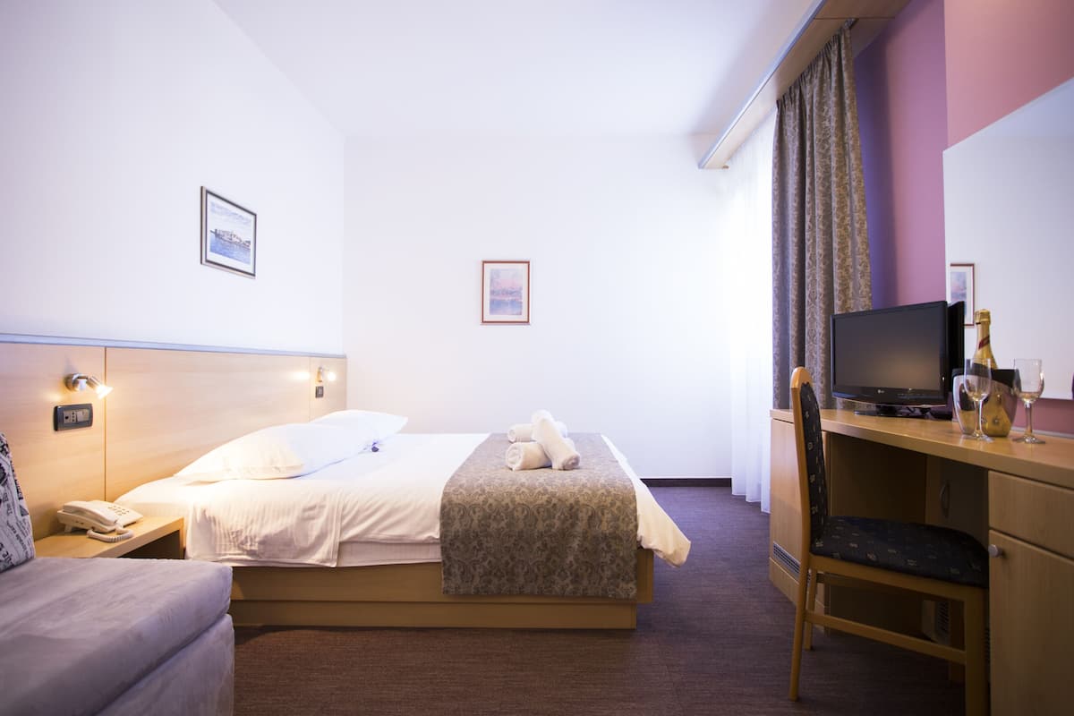 A hotel room with a bed, a desk with a computer, a chair, a telephone, and two framed pictures on the wall.