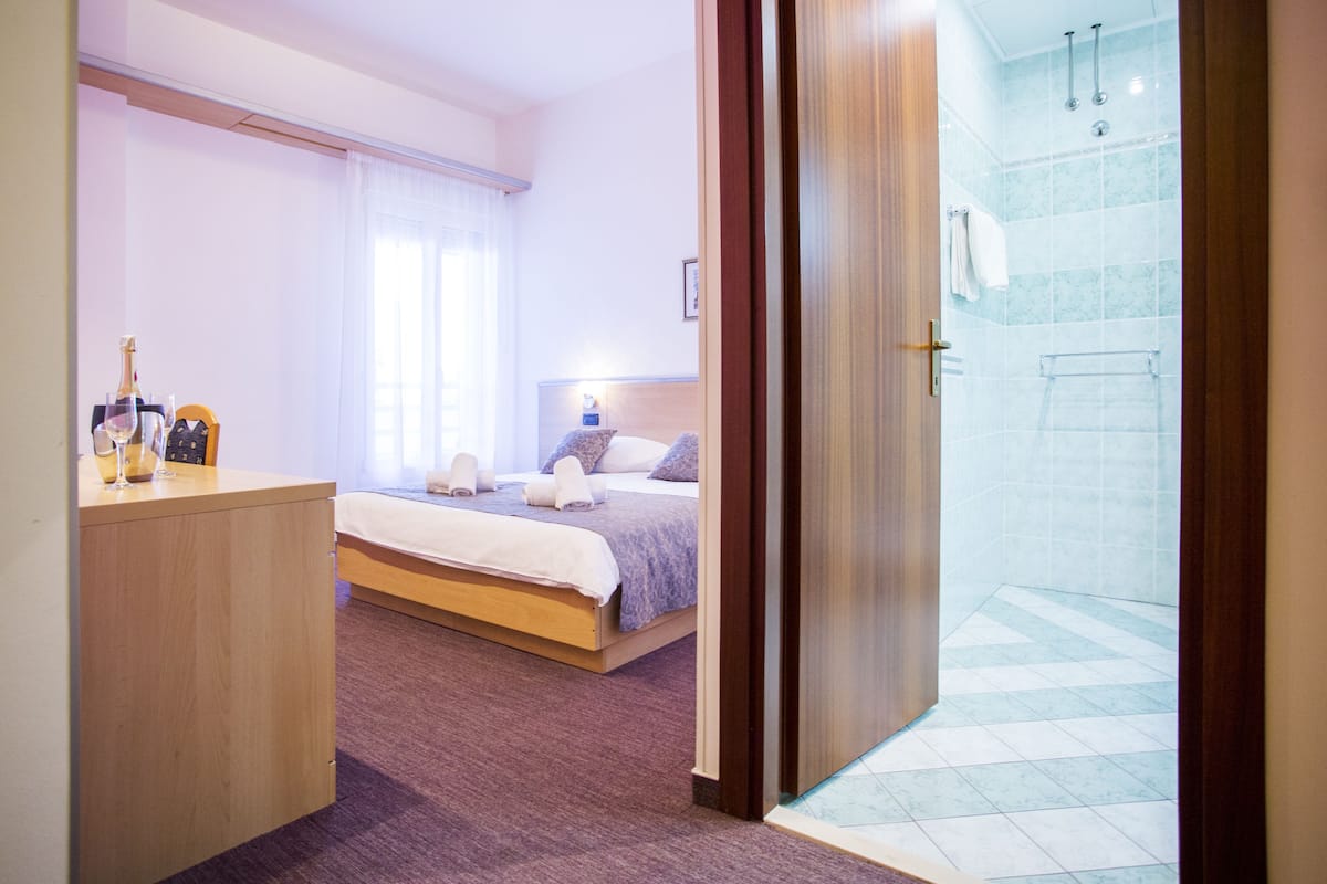 A hotel room with a bed, a bedside table with a bottle and glass, a shower area with a glass door, and a bathroom with a towel rack.
