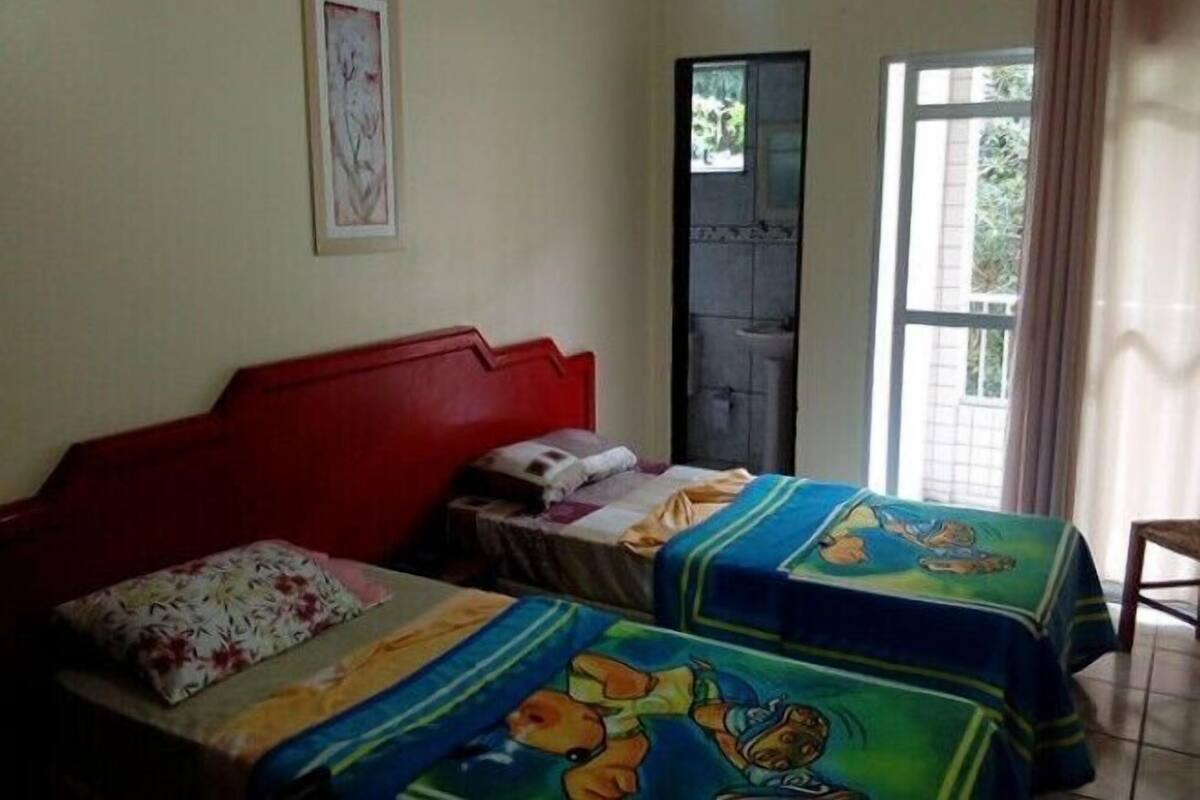 Ein Schlafzimmer mit rotem Kopfteil, zwei Betten mit bunter Bettwäsche, einem kleinen Tisch und einem Fenster mit Vorhängen.