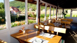 A dining area with wooden tables and benches, large windows with a view of a garden and houses, and a sunny atmosphere. - Pension Perisic (Plitvicka Jezera)