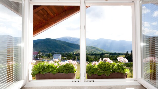 A balcony with wooden flooring, potted plants, and a view of mountains and a village. - Pension Perisic (Plitvicka Jezera)