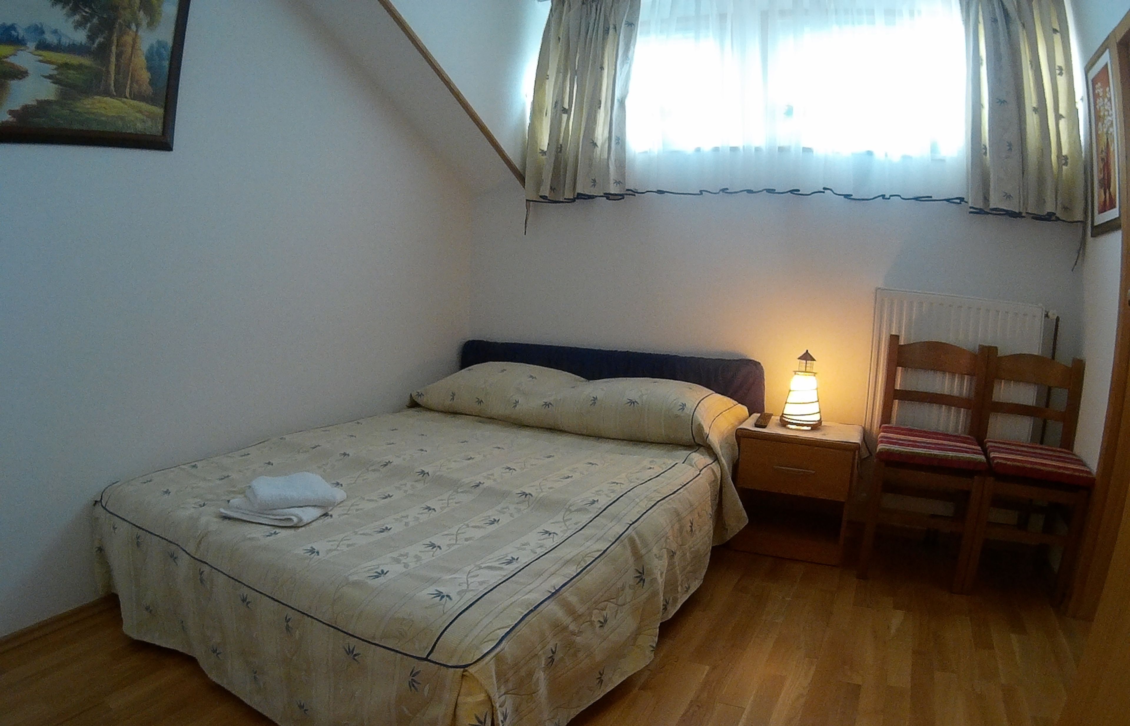 Un dormitorio con cama, mesita de noche, una silla y una ventana con cortinas.