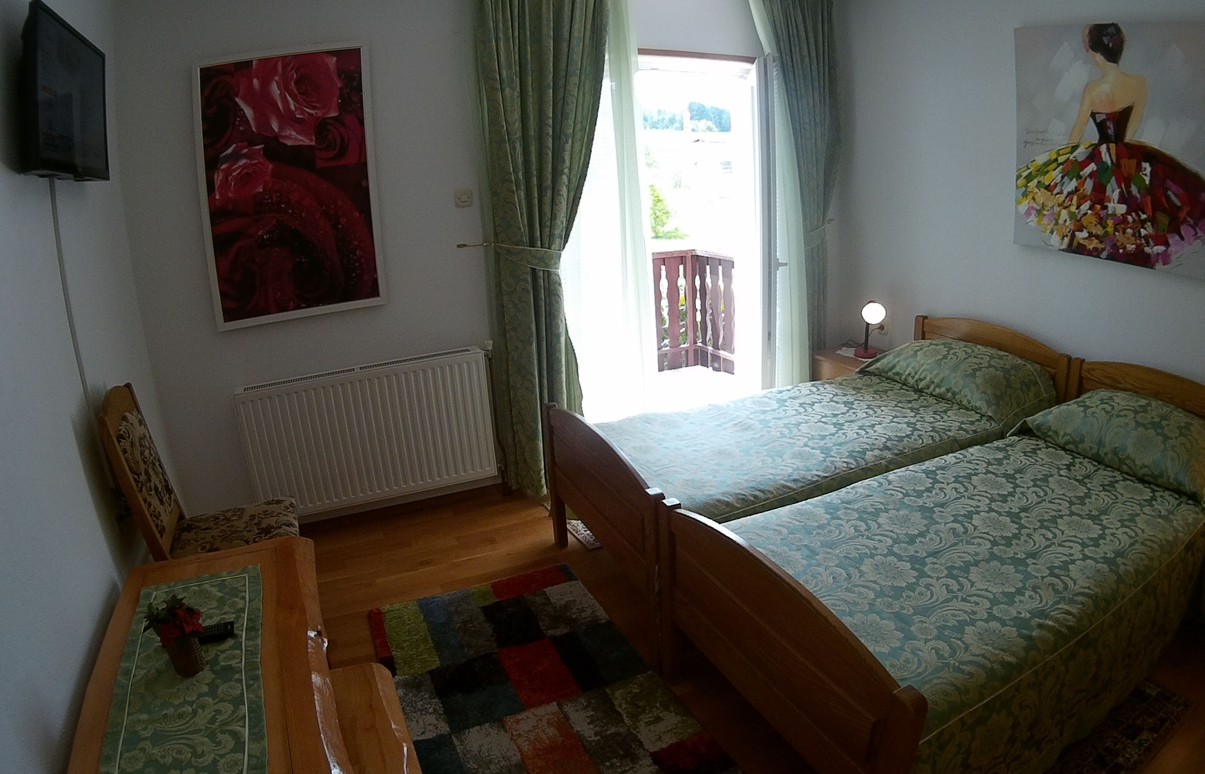 Um quarto com uma cama, uma mesa de cabeceira de madeira, uma mesa com abajur, um radiador, uma janela com cortinas e um quadro na parede.