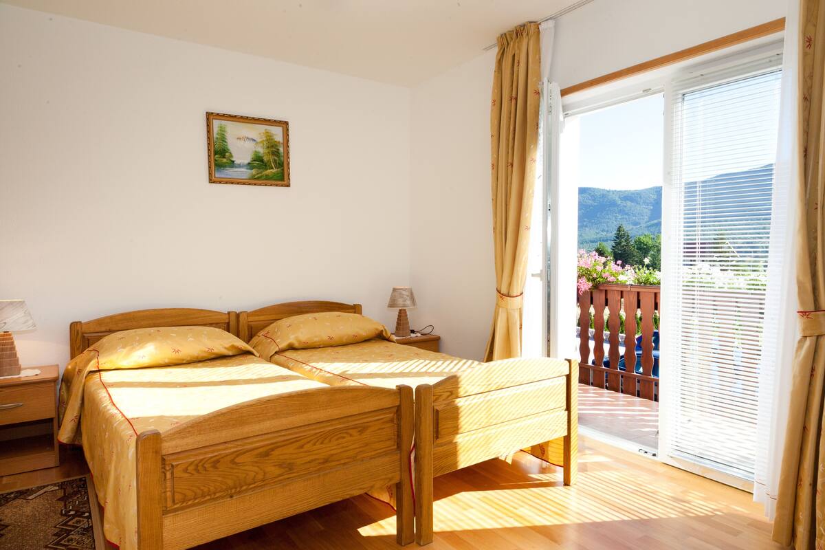 Una cama doble con estructura de madera y cabecera, una mesita de noche con lámpara, y una ventana con cortinas.