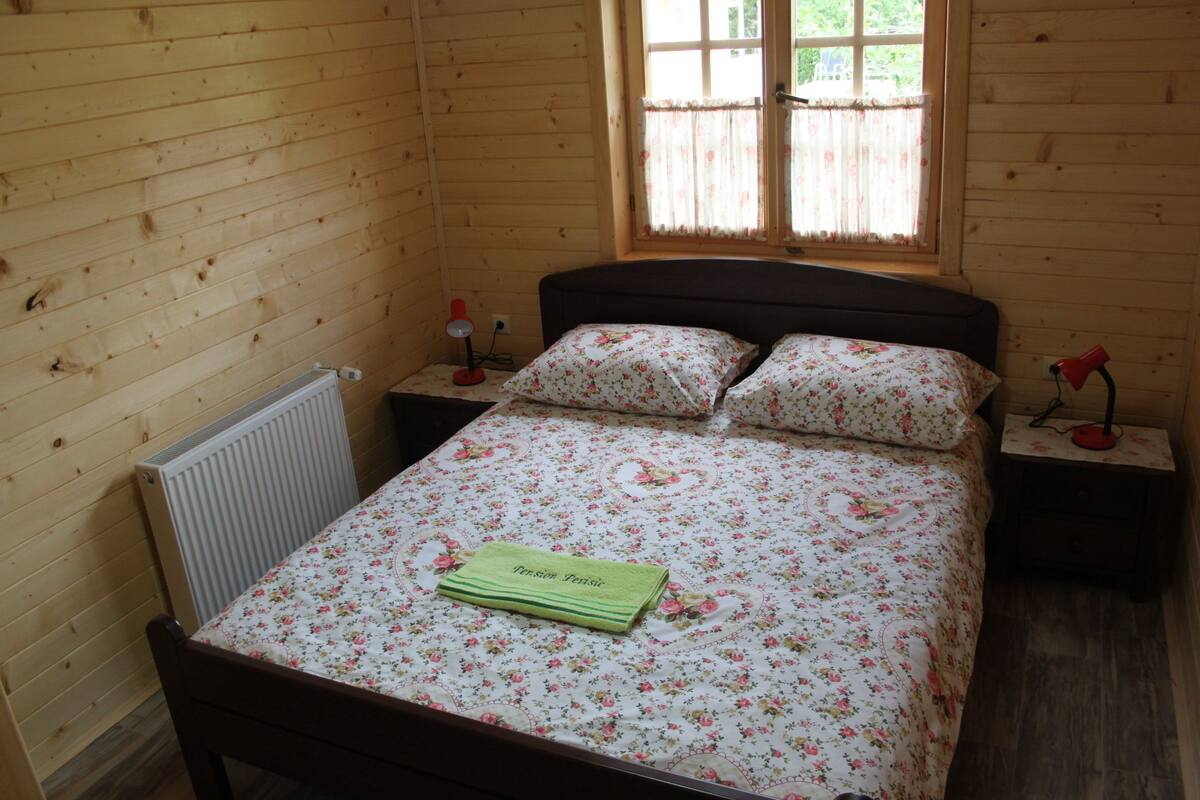 Un dormitorio de cabaña de madera con una cama, dos mesitas de noche, un ventanal con cortinas y un radiador.