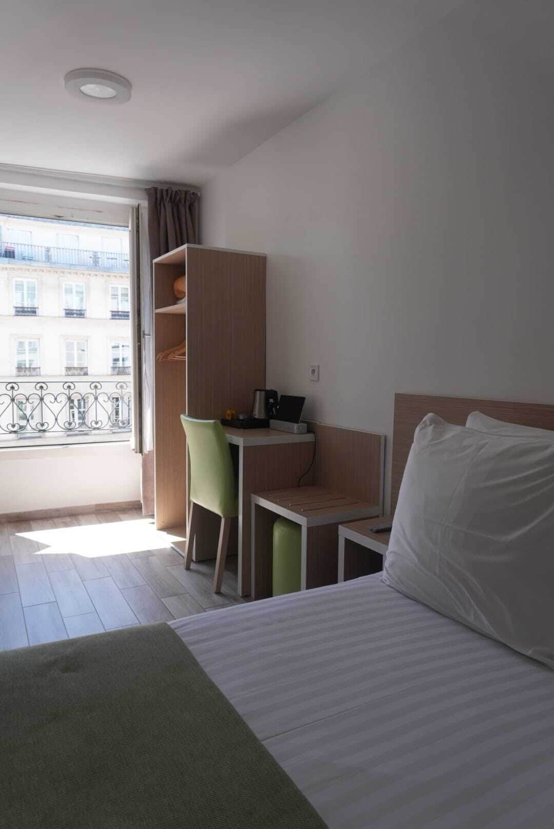 Habitación de hotel con cama, escritorio, silla y armario. Se ve una ventana con vistas a edificios.