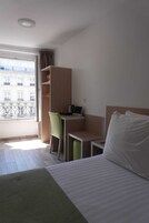 Camera d'albergo con un letto, una scrivania, una sedia e un armadio. Si vede una finestra con vista sugli edifici.