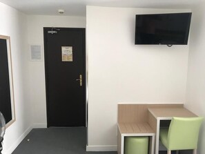 Una habitación pequeña con una puerta negra, un televisor blanco montado en la pared, una mesa de madera y dos sillas verdes.