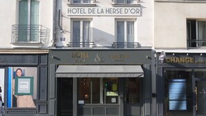 Un edificio con un cartel que dice 'HOTEL DE LA HERSE D'OR' y una tienda con el rótulo 'HOTEL & CONSIGNE A BAGAGES', un código QR y una pantalla digital.