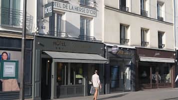 Un hôtel avec une enseigne indiquant « HÔTEL DE LA HERSE D’OR » et une devanture portant les inscriptions « HÔTEL ET DÉPÔT-BAGAGES » et « CHANGE ».