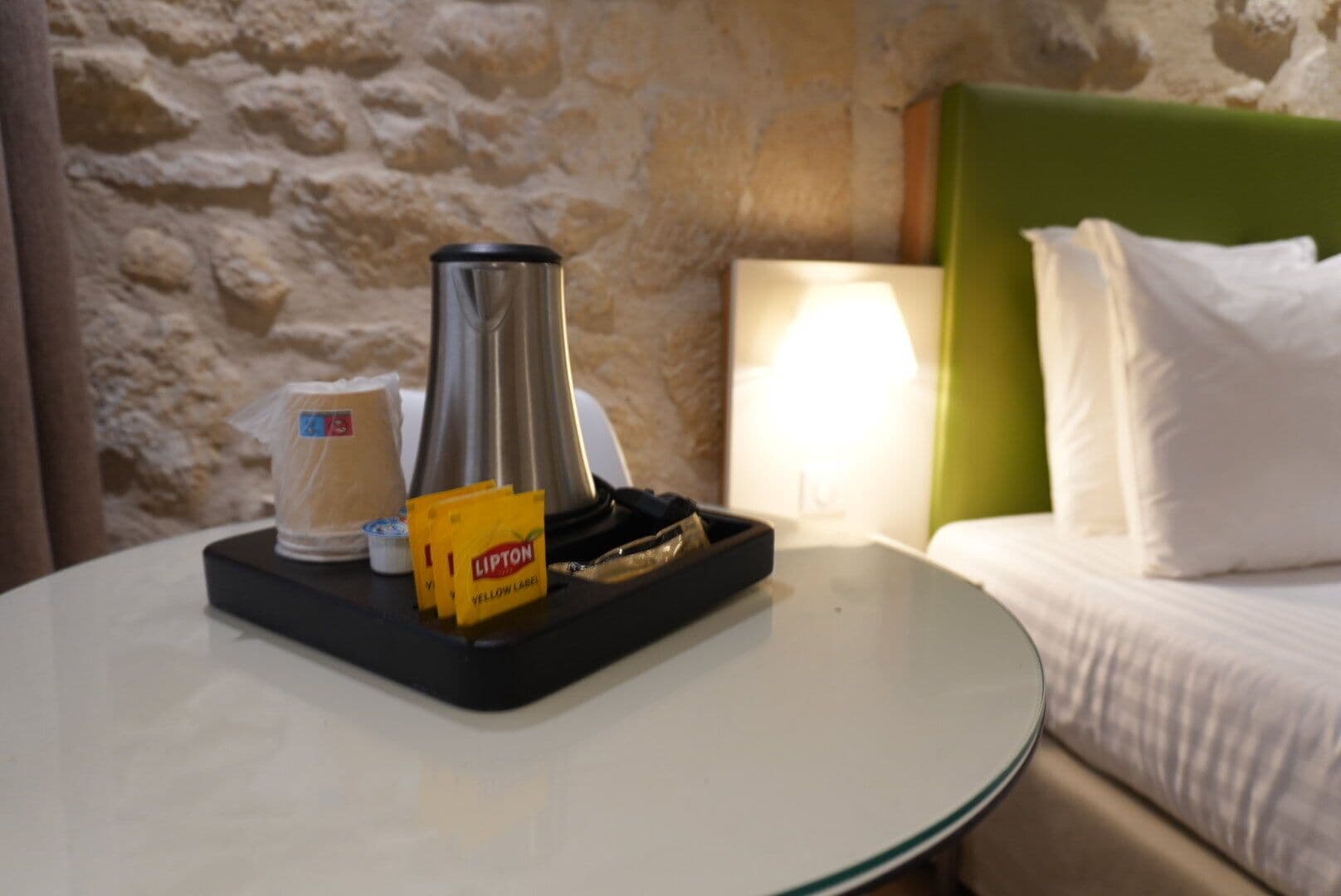 A hotel room with a bed, a nightstand, a lamp, a coffee maker, a cup, and a tray with Lipton tea bags.