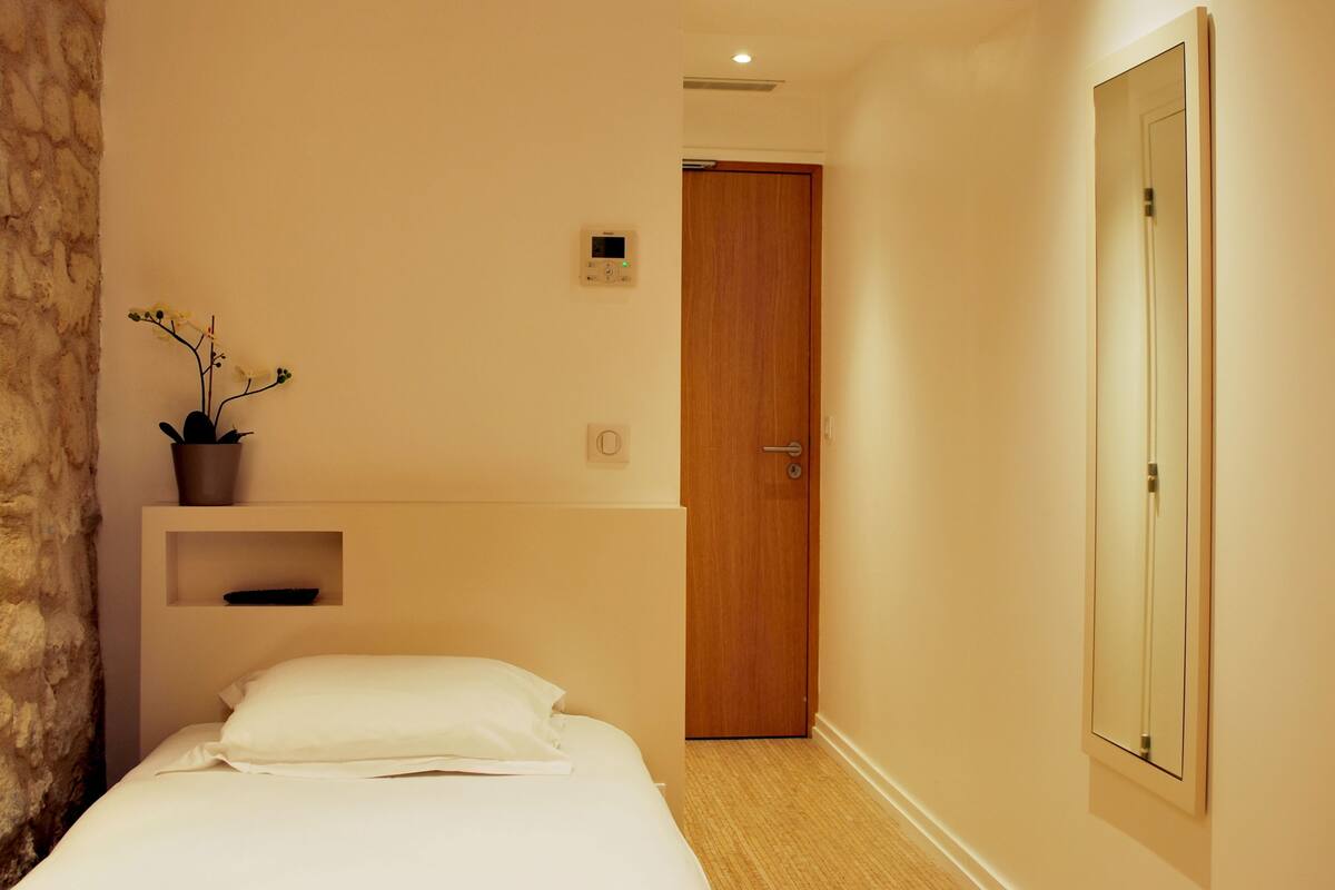 Una habitación de hotel con cama, una pequeña estantería con una planta, una puerta y una lámpara fijada a la pared.