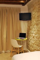A hotel room with a bed, a desk with a laptop, a chair, a television mounted on a stone wall, and beige curtains.