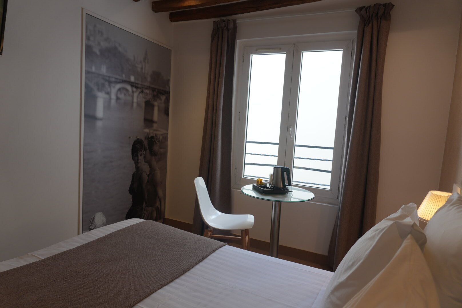 A hotel room with a bed, a small table with a kettle, a chair, and a window with curtains.