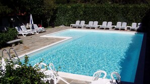 A rectangular swimming pool with multiple lounge chairs and a table on one side, surrounded by greenery.