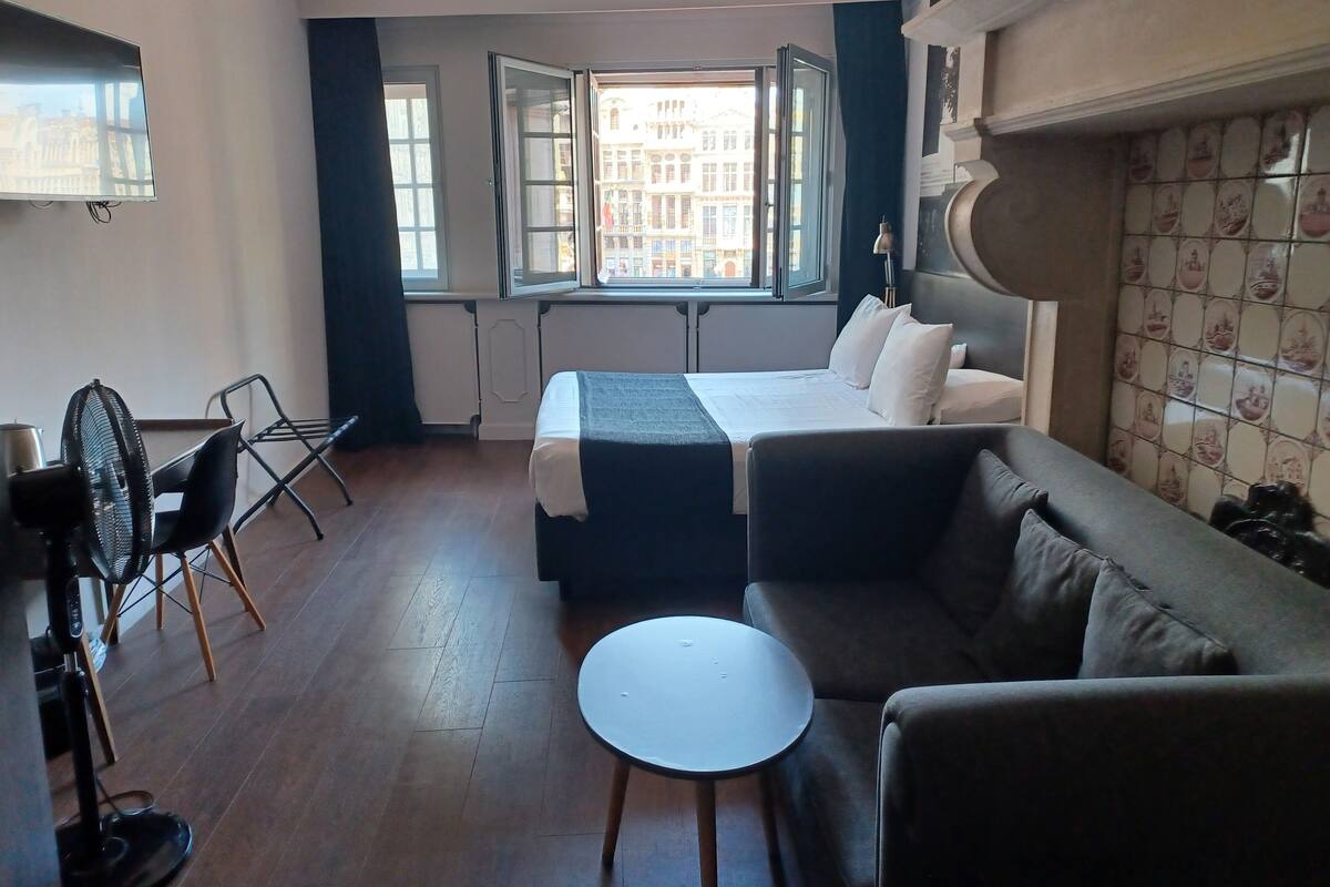 Una habitación de hotel con cama, sofá, una pequeña mesa redonda, una silla y vistas a la ciudad a través de grandes ventanales.