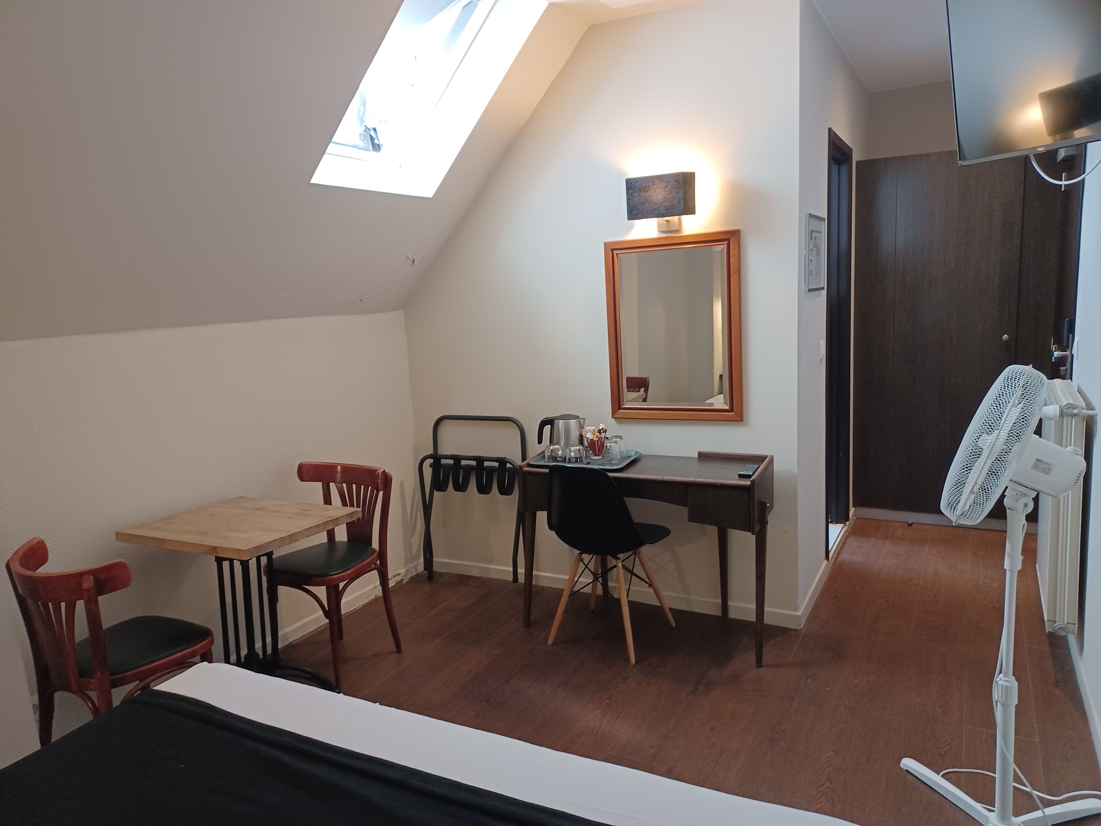Una habitación pequeña y bien iluminada con un escritorio, dos sillas, una cama, un espejo y un ventilador de techo.