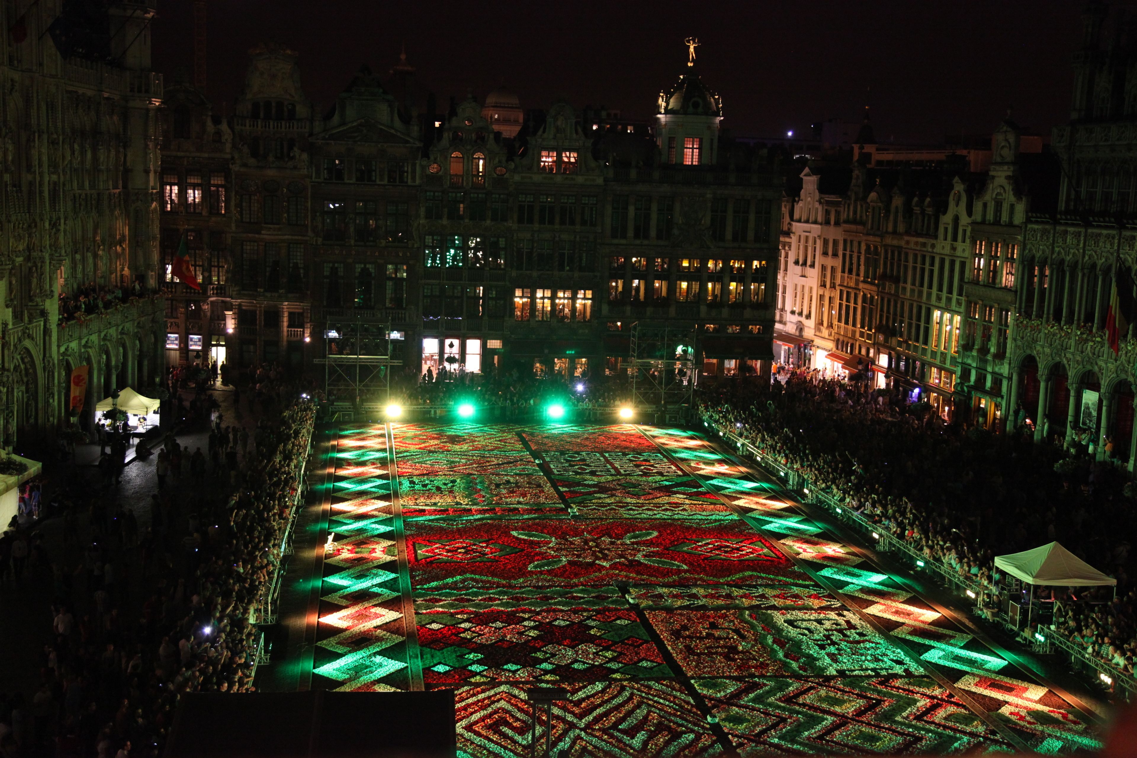 Una plaza histórica con edificios ornamentados y un gran tapiz floral iluminado.