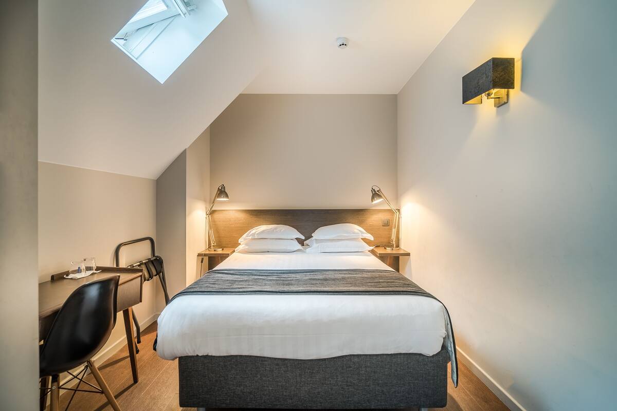 Una habitación de hotel moderna con tragaluz, cama con ropa blanca, escritorio con silla y lámparas fijadas a la pared.